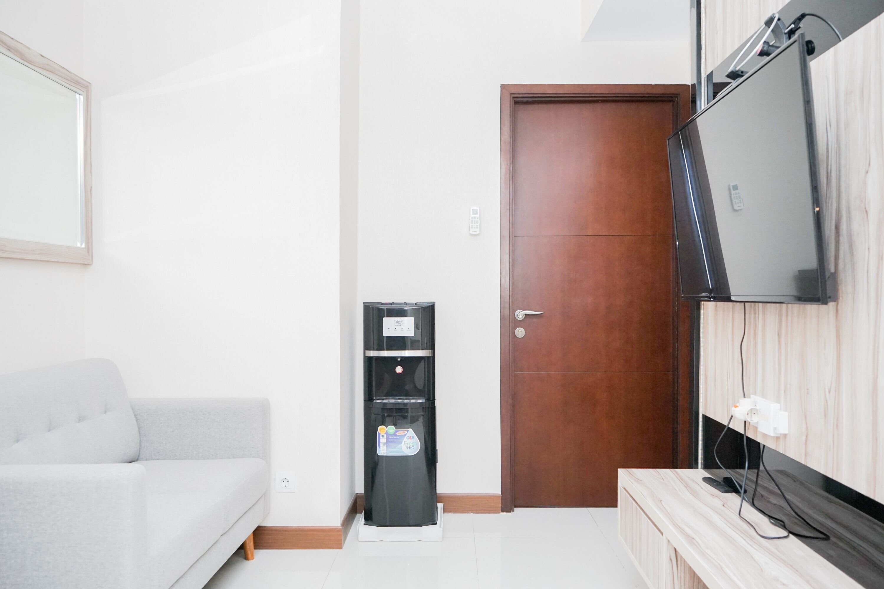 Un salon moderne avec un canapé gris, un distributeur d’eau noir, une porte en bois et un téléviseur à écran plat fixé au mur.