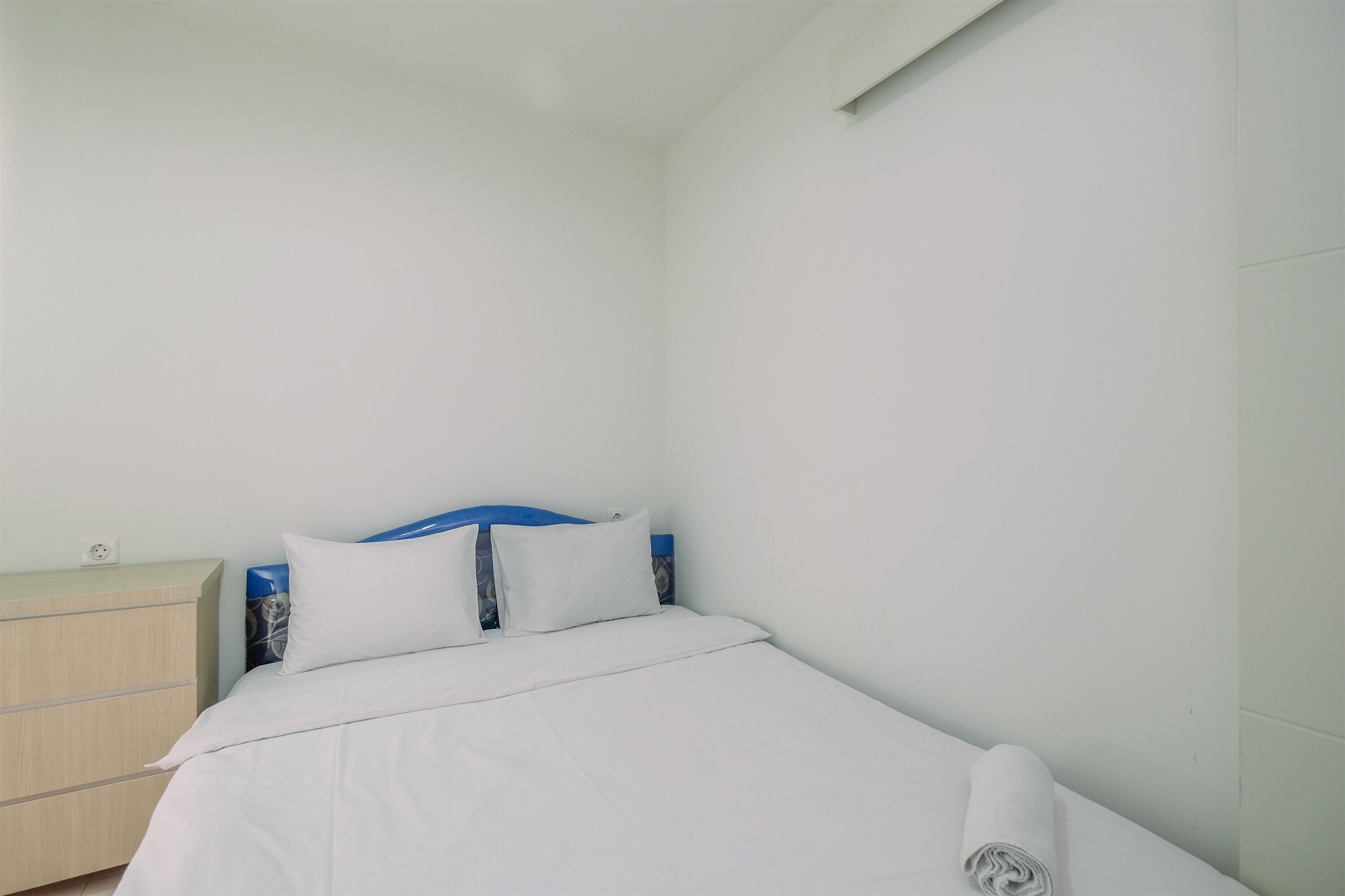 A neatly made bed with white linens and pillows, a wooden nightstand with a power outlet, and a blue headboard.