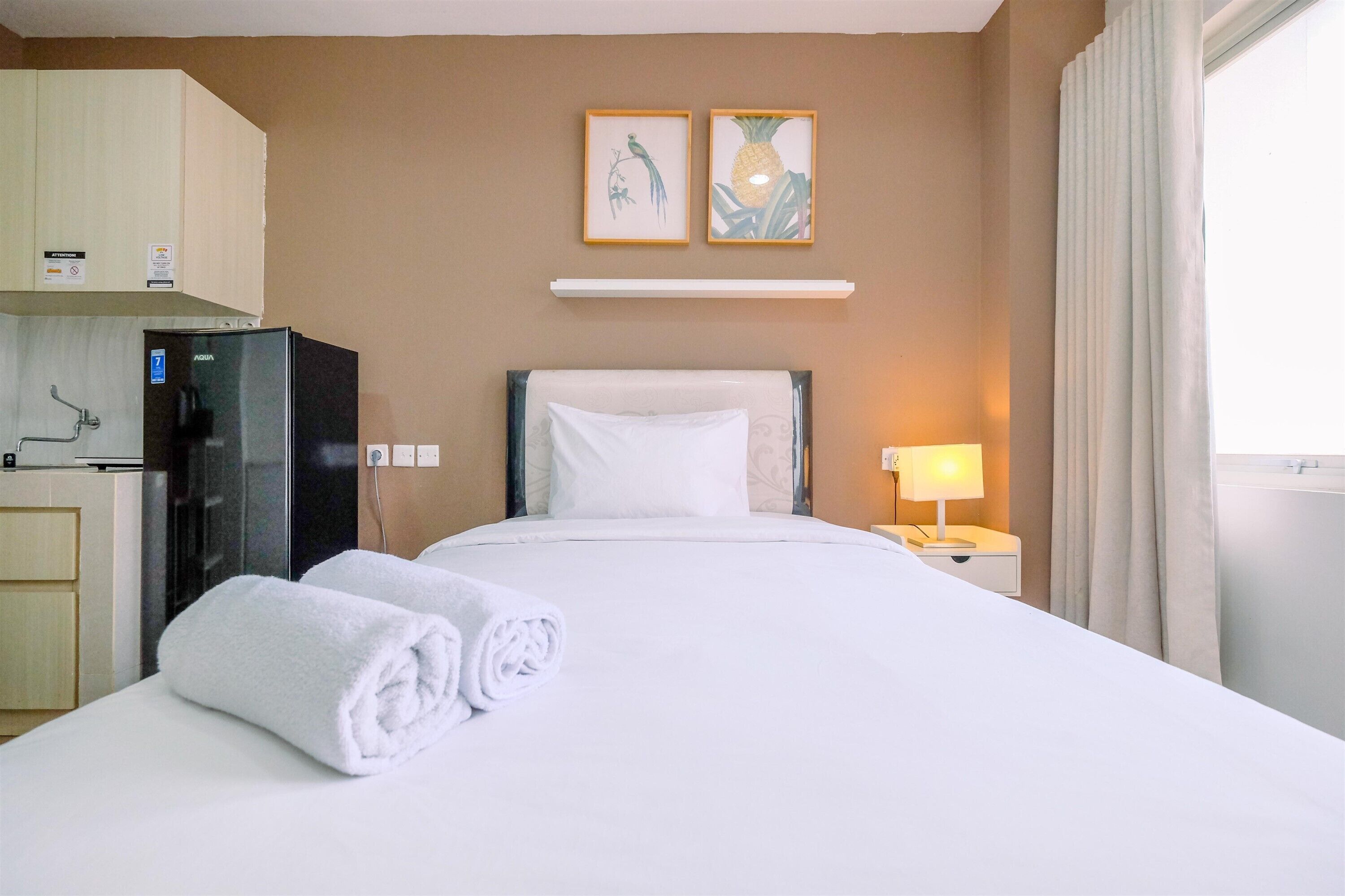 Een hotelkamer met een bed, een nachtkastje met een lamp, een koelkast, en een wandplank met twee ingelijste foto's.