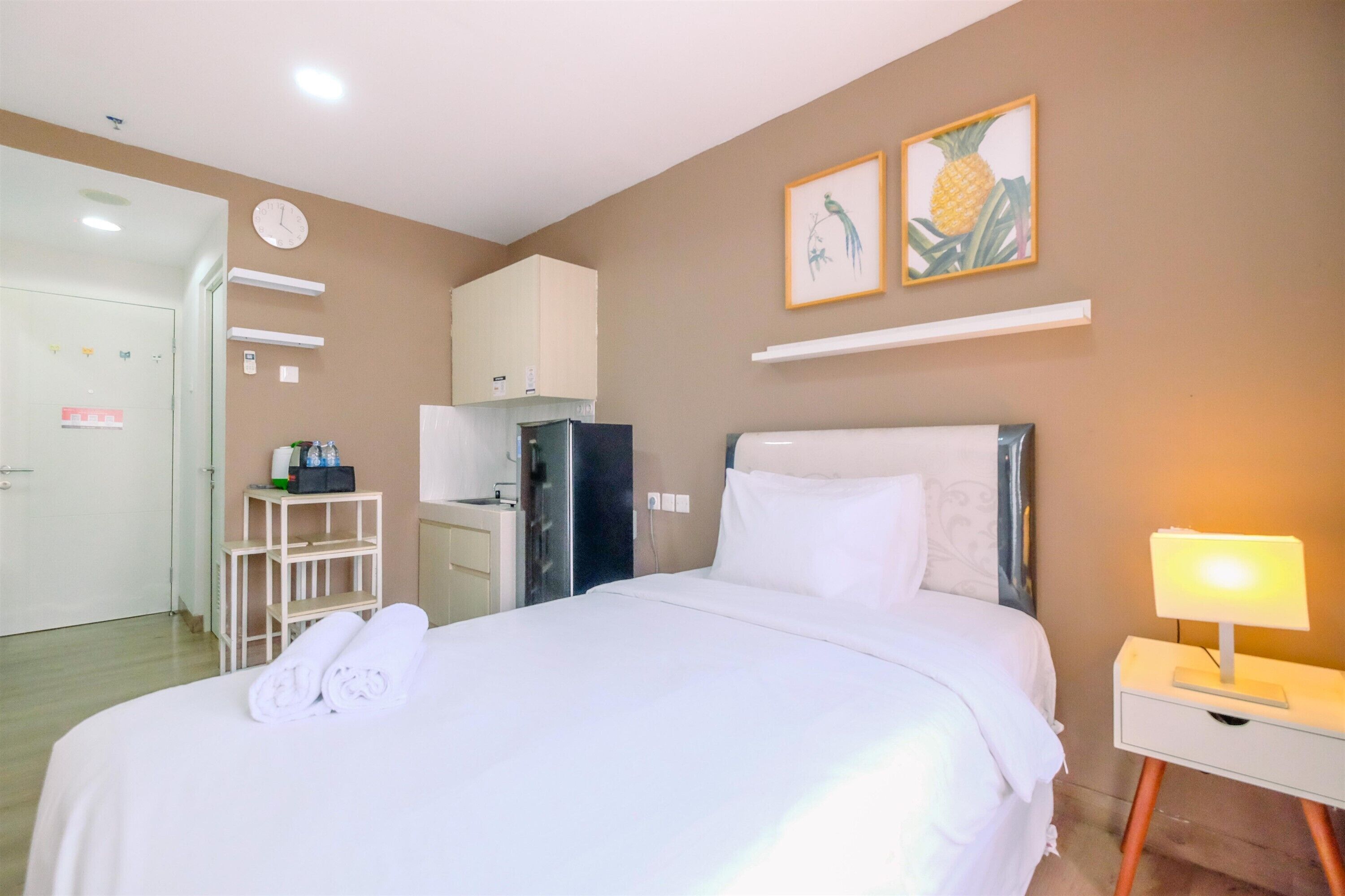 A hotel room with a bed, a nightstand with a lamp, a refrigerator, a small kitchen area, and two wall-mounted artworks.