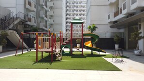 A playground with a slide and swings, surrounded by a green lawn, in an urban residential area with high-rise buildings in the background. - Relax 1BR Apartment at Sentul Tower (Citaringgul)