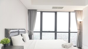 A modern hotel room with a large bed, a bedside table with a plant, and a view of the city through large windows. - Cozy and Elegant Studio The Smith Alam Sutera Apartment (Tangerang)