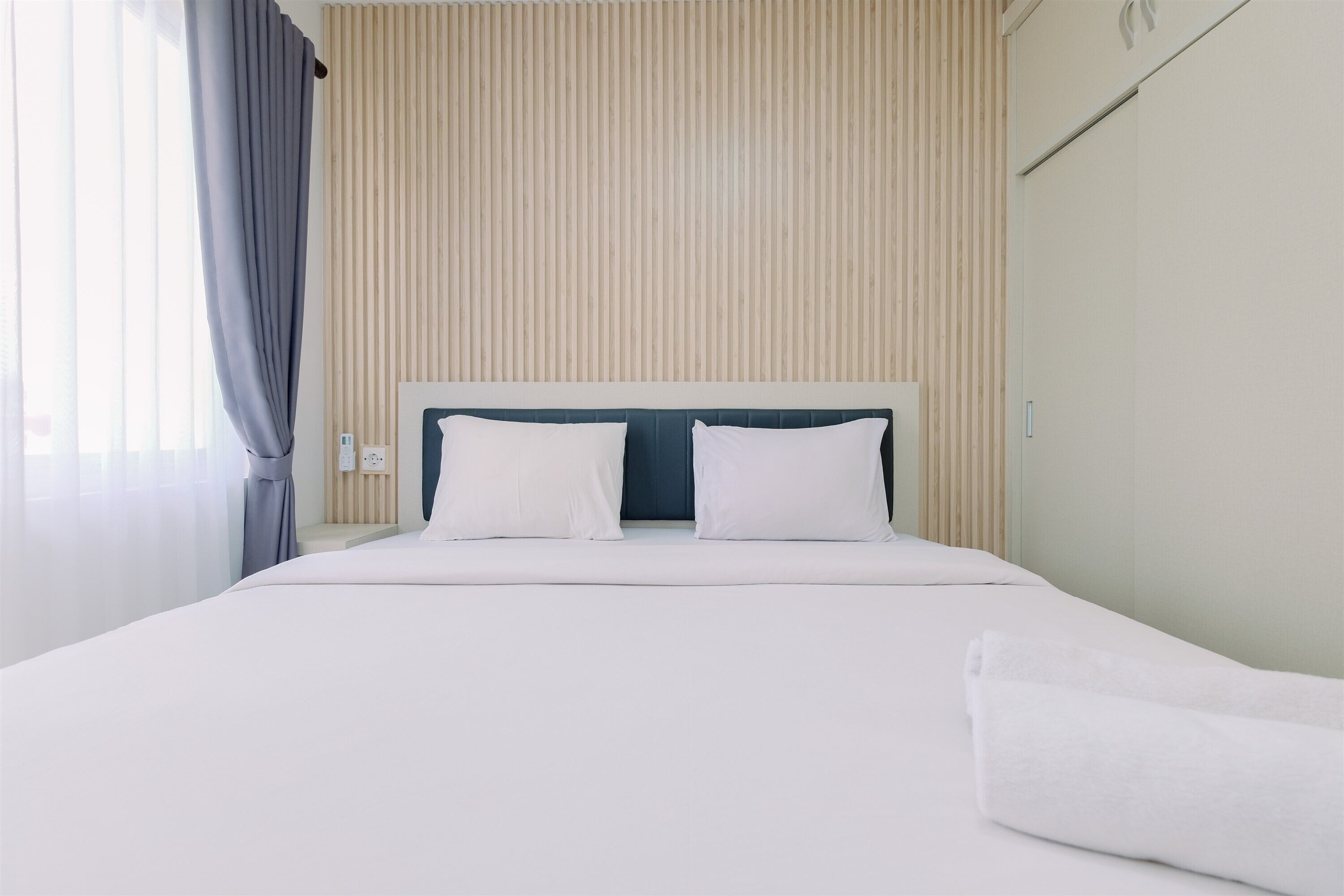 A neatly made bed with white linens and pillows, a wooden slat wall, and a window with curtains.