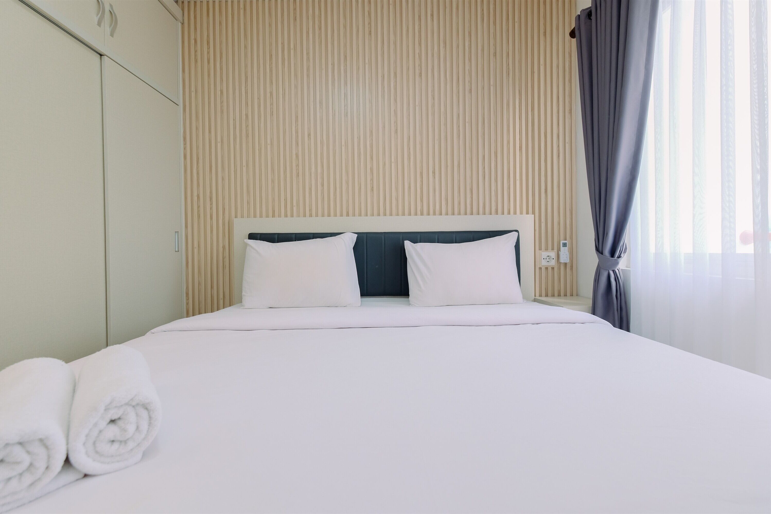 A neatly made bed with white linens and pillows, a wooden slat wall, and a window with curtains.