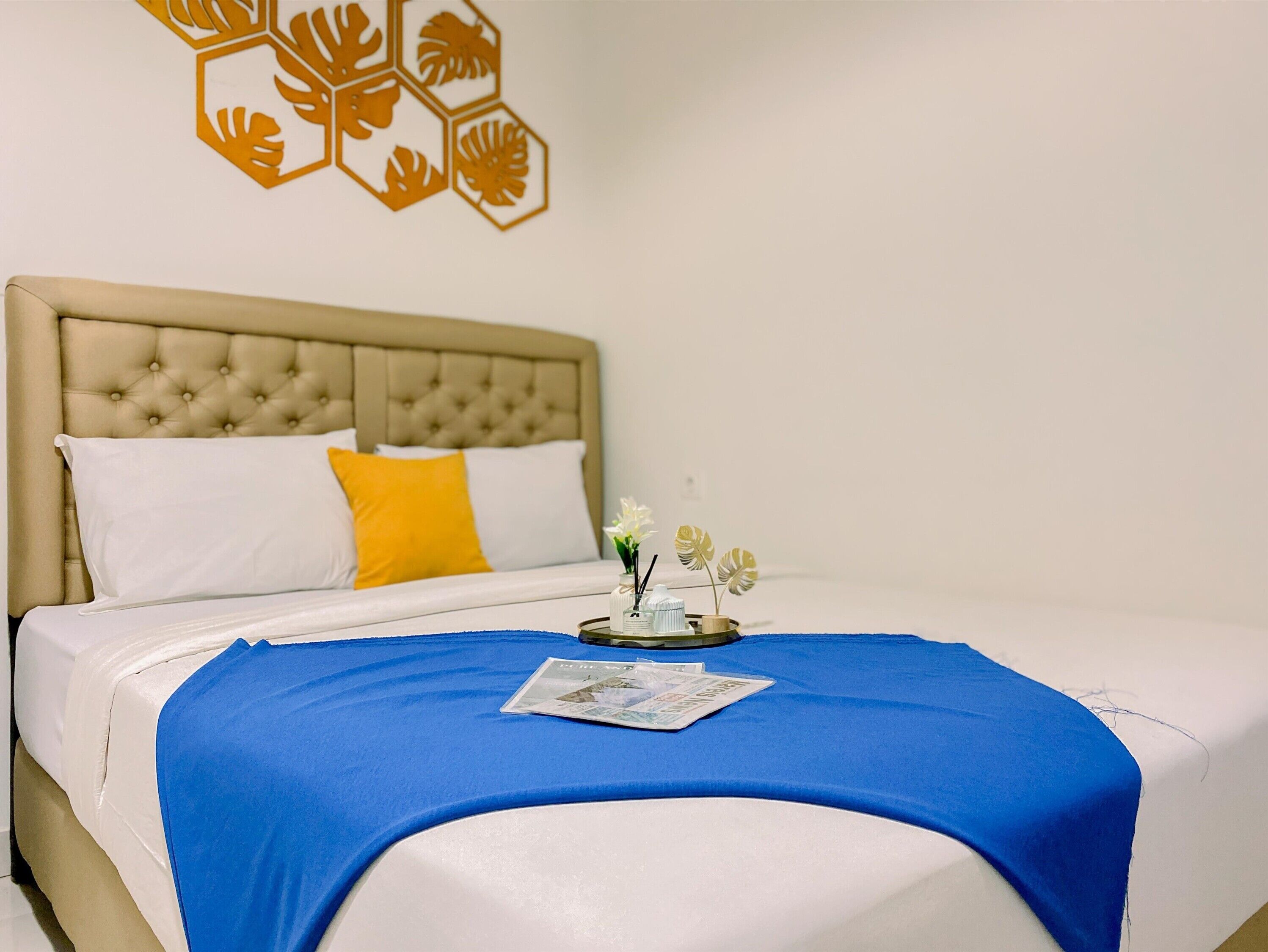 Una cama bien hecha con una manta azul, un almohadón blanco y un almohadón amarillo. Hay una mesita con un florero y una revista.