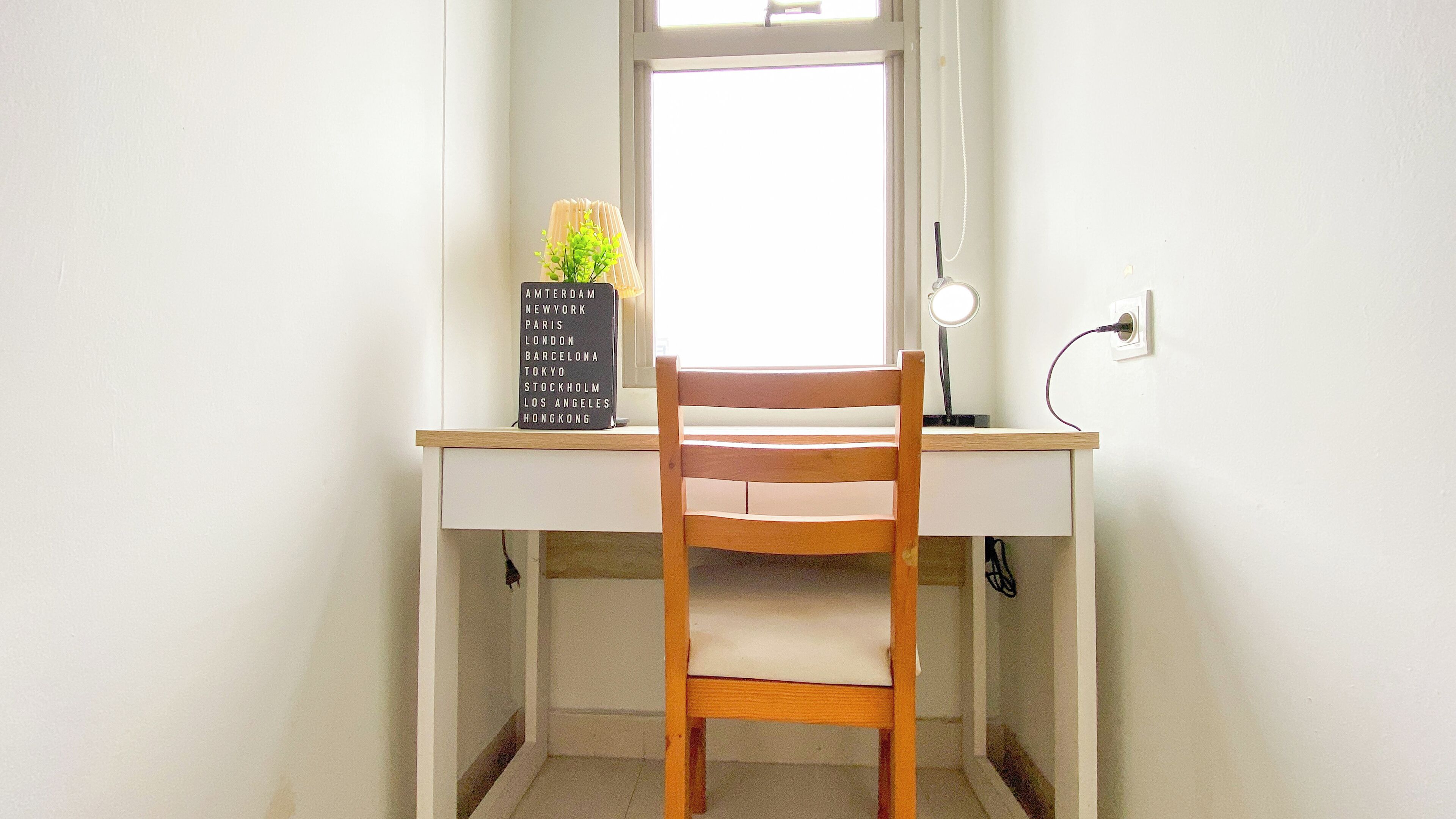A wooden chair facing a white desk with a window. A black sign with city names is on the desk.