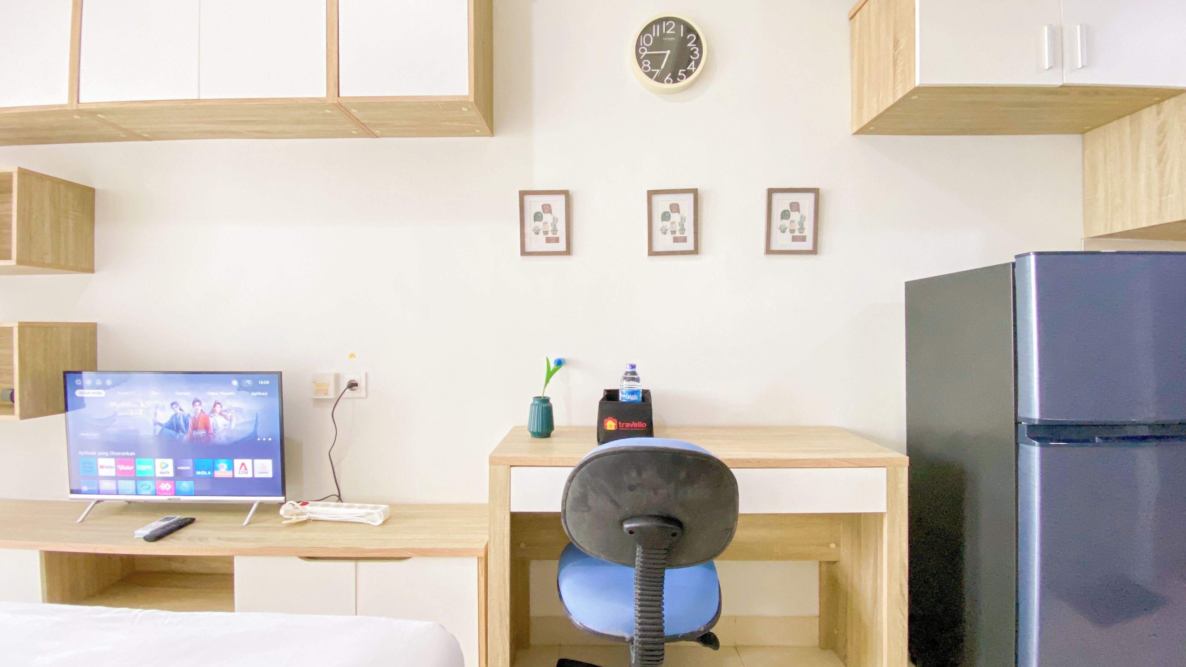 A compact hotel room with a wooden desk, a small chair, a television, a refrigerator, and a wall clock.
