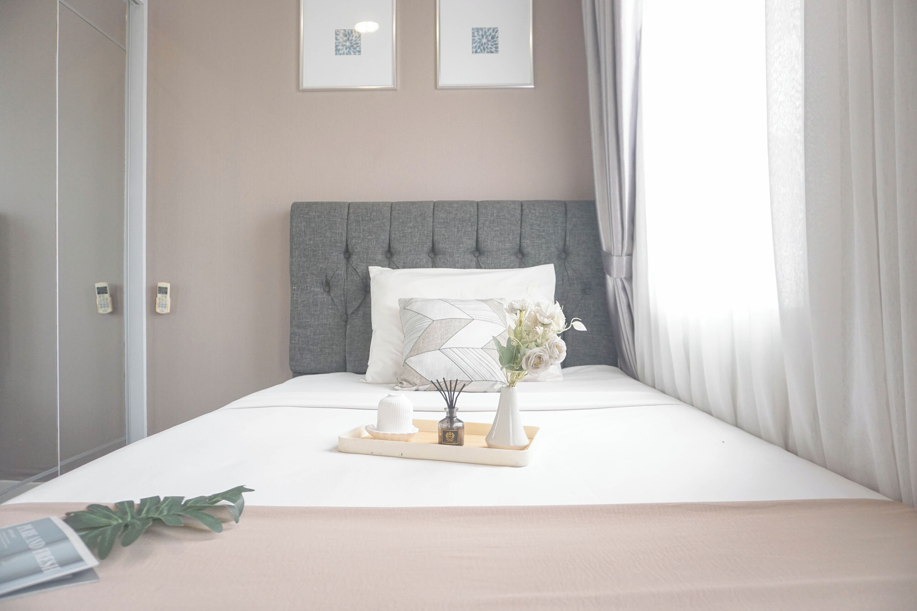 A neatly made bed with a grey headboard, a white pillow, and a tray with a vase of flowers and a candle.