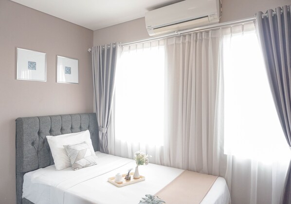 A neatly made bed with a grey headboard, a small bedside table with a vase of flowers, and a window with sheer curtains. - Cozy and Spacious 2BR at Sentul Tower Apartment (Citaringgul)