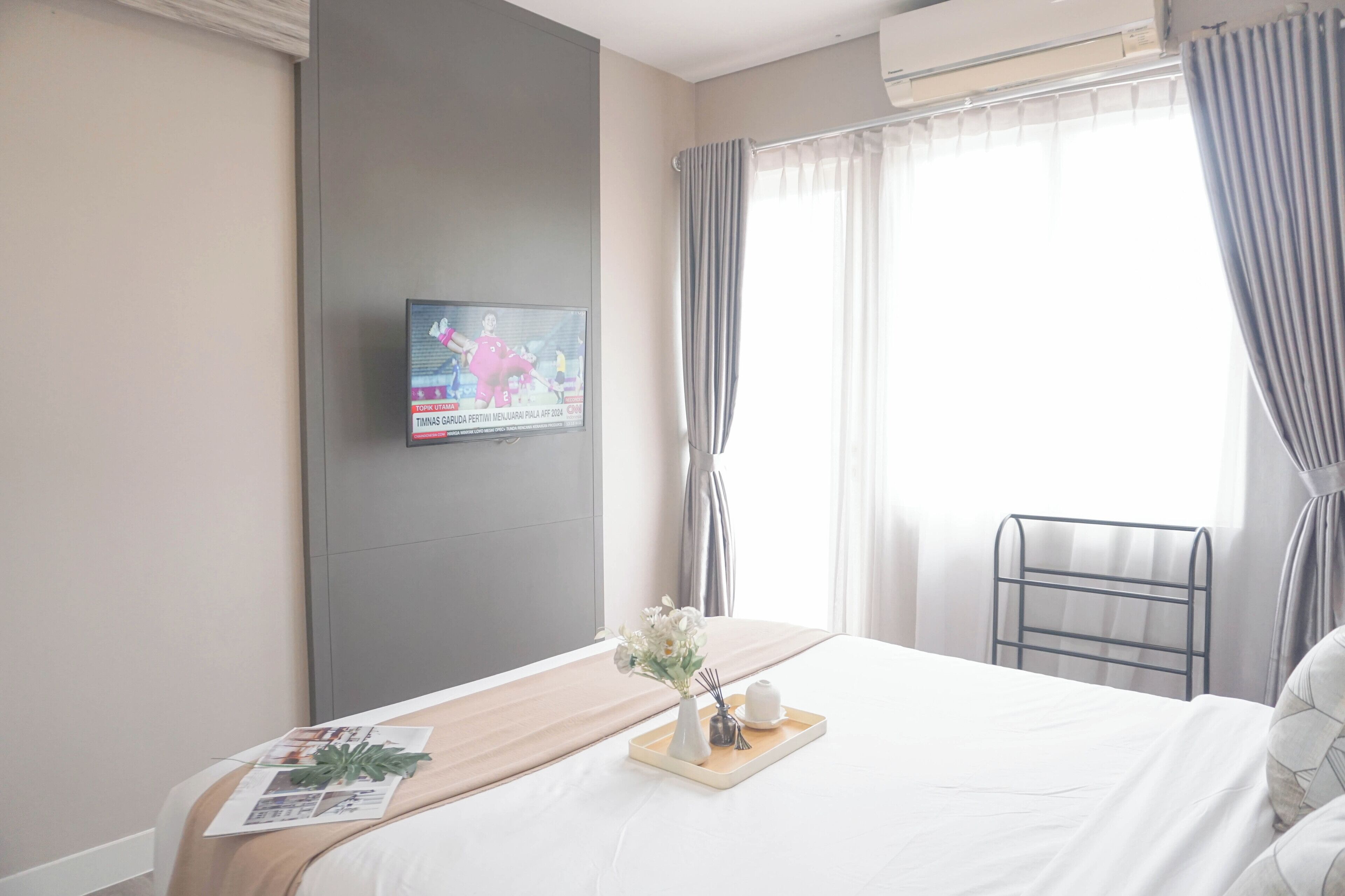 Ein Hotelzimmer mit einem Bett, einem an der Wand montierten Fernseher, einem Fenster mit Vorhängen und einem kleinen Tisch mit Tablett.