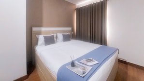 Habitación de hotel con una cama grande, cabecera, ventana con cortinas y mesita de noche con lámpara y revista.