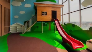 Zona de juegos infantiles con un tobogán, una casita de madera y vistas amplias a través de grandes ventanales.