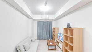 A modern living room with a sofa, a coffee table, a TV on a wooden stand, and a large window with curtains. - Spacious and Homey 2BR at Podomoro City Deli Medan Apartment (Medan)
