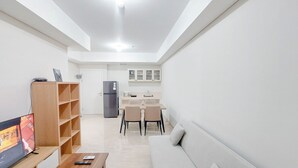 A modern living room with a sofa, a coffee table, and a TV. There is a wooden bookshelf and a kitchen area with a fridge and cabinets. - Spacious and Homey 2BR at Podomoro City Deli Medan Apartment (Medan)