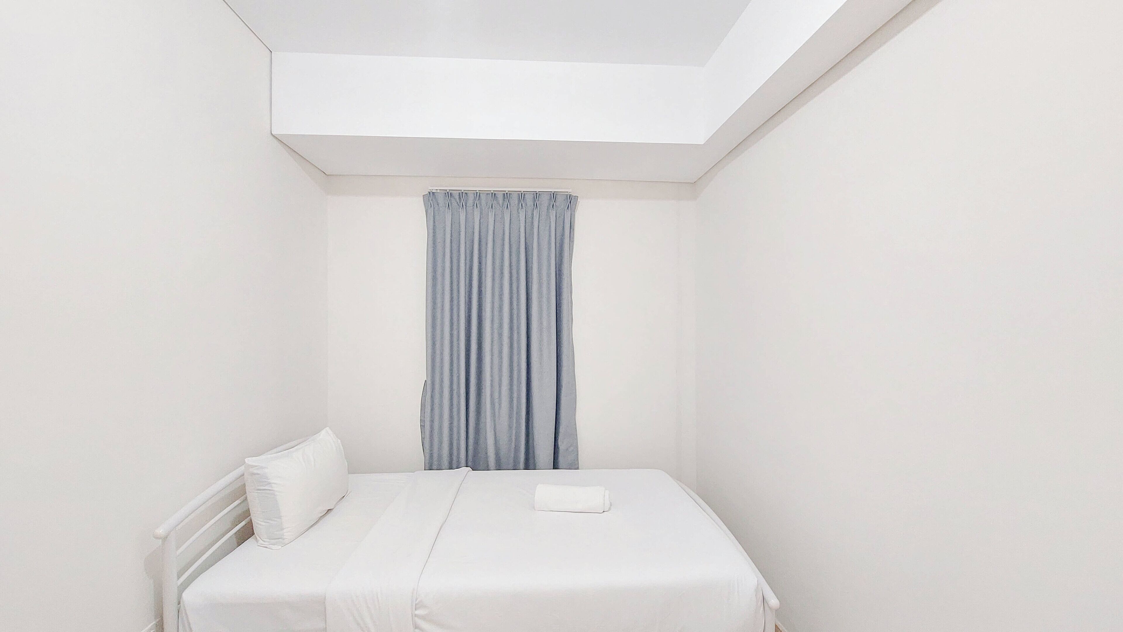 Una habitación pequeña y blanca con una cama individual, techo inclinado y ventana con cortinas azules.