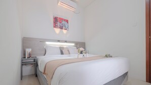 A modern hotel room with a large bed, a bedside table with a lamp, and a wall-mounted air conditioner. - Well Designed and Warm 2BR Apartment at Vittoria Residence (Jakarta)