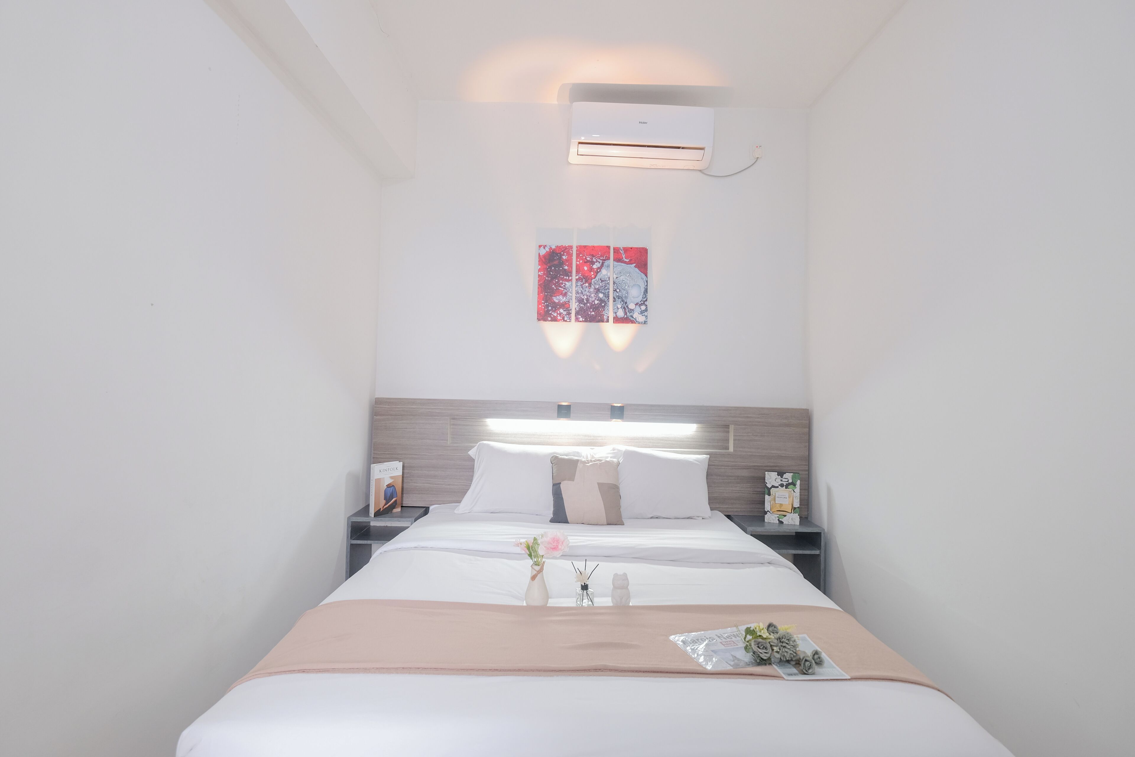 A modern hotel room with a neatly made bed, a bedside table with a lamp, a wall-mounted air conditioner, and a window with a view of the city.