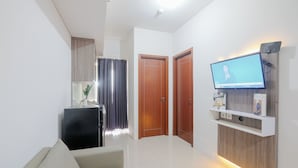 A modern living room with a sofa, a flat-screen TV mounted on a wooden wall, a small cabinet, and a door leading to another room. - Cozy and Sweet 2BR Apartment at Vittoria Residence (Jakarta)