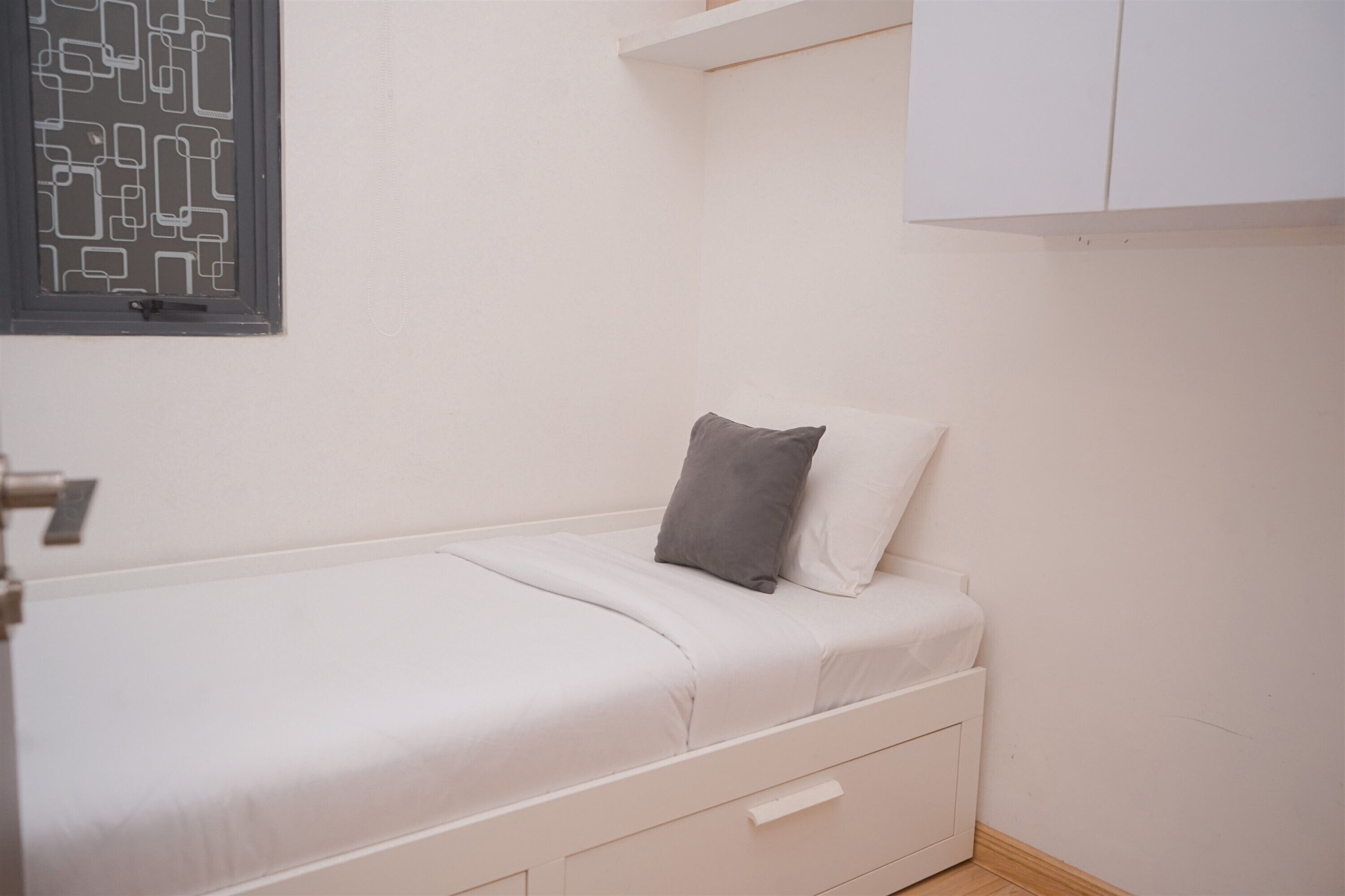 Habitación pequeña con cama individual, almohada gris y un mueble blanco sobre la cama. Se observa también un radiador fijado a la pared.