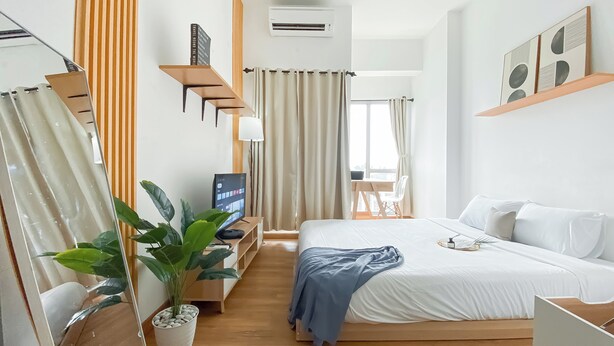 Ein modernes Hotelzimmer mit einem großen Bett, einem Schreibtisch mit Computer, einer Zimmerpflanze und einem Vorhangfenster.