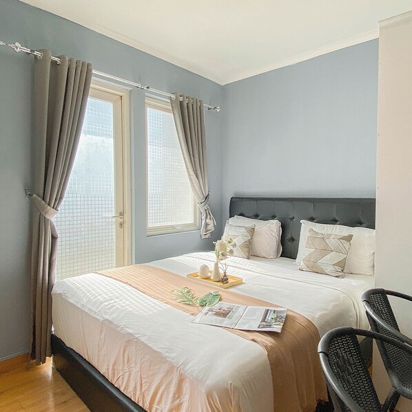 A bedroom with a large bed, two windows with curtains, a bedside table with a vase, and a chair. - Warm and Cozy Studio at Sentul Tower Apartment (Citaringgul)
