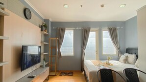 Een kleine hotelkamer met een bed, een bureau met een televisie, twee stoelen, een trapje en een raam met gordijnen.
