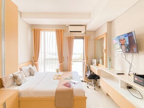 A hotel room with a bed, a desk, a chair, a TV, and a window with curtains. - Comfort Living Studio at Monroe Tower Apartment (Cikarang)