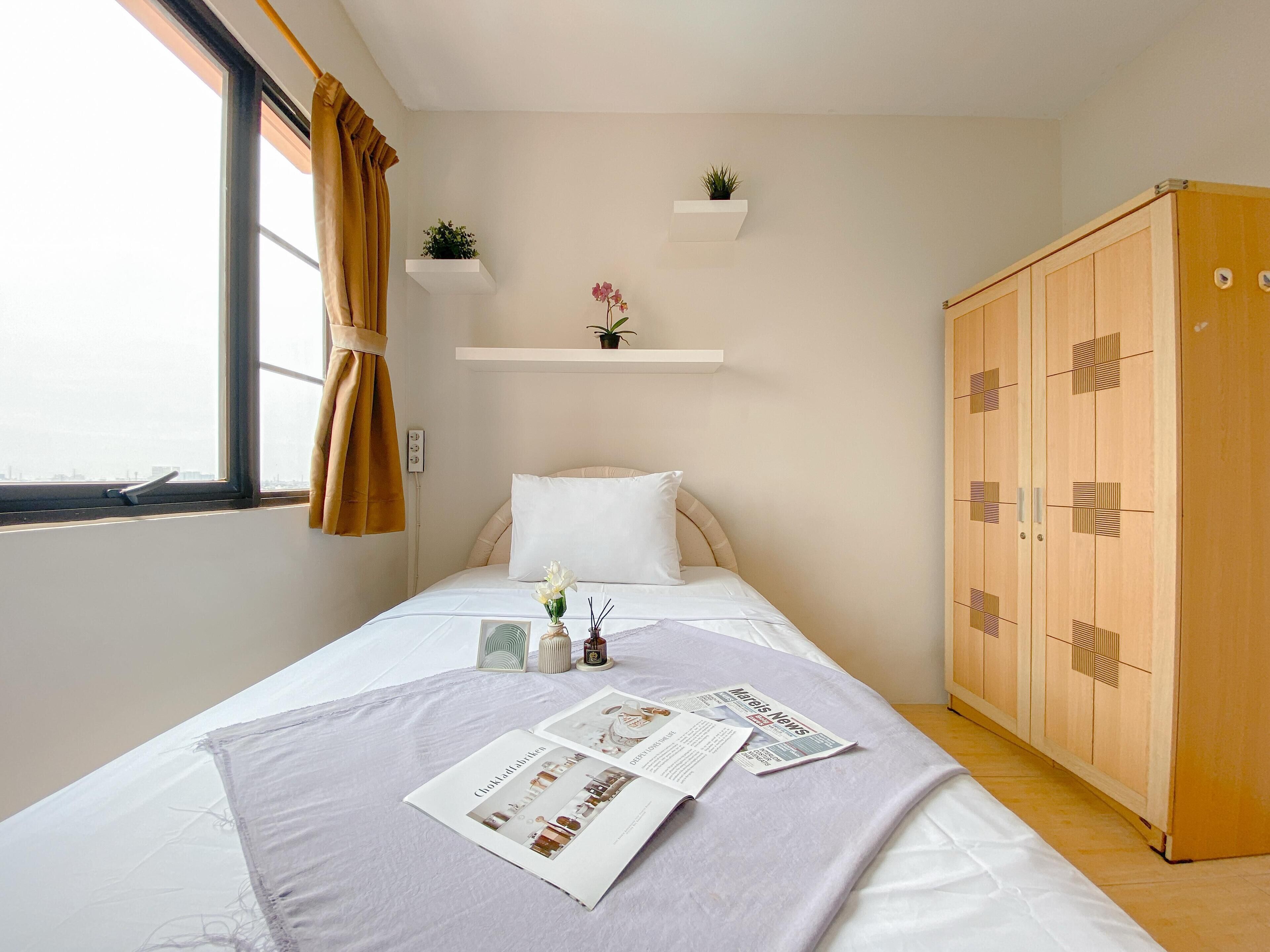 A neatly made bed with a magazine and a small plant on the bedside table, a wardrobe with a geometric design, and a window with curtains.
