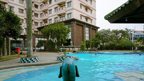 Ein Schwimmbecken im Vordergrund, geschmückt mit einer dekorativen Skulptur, umgeben von einer angelegten Grünfläche mit Bäumen und einem mehrstöckigen Gebäude im Hintergrund.