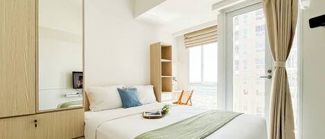 A hotel room with a bed, a wooden wardrobe, a small desk, and a window with curtains.