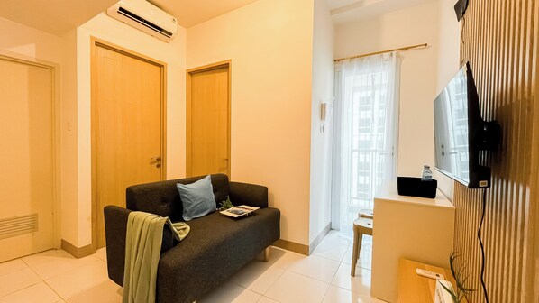 A modern living room with a dark sofa, a small table, a TV mounted on the wall, and a window with blinds.
