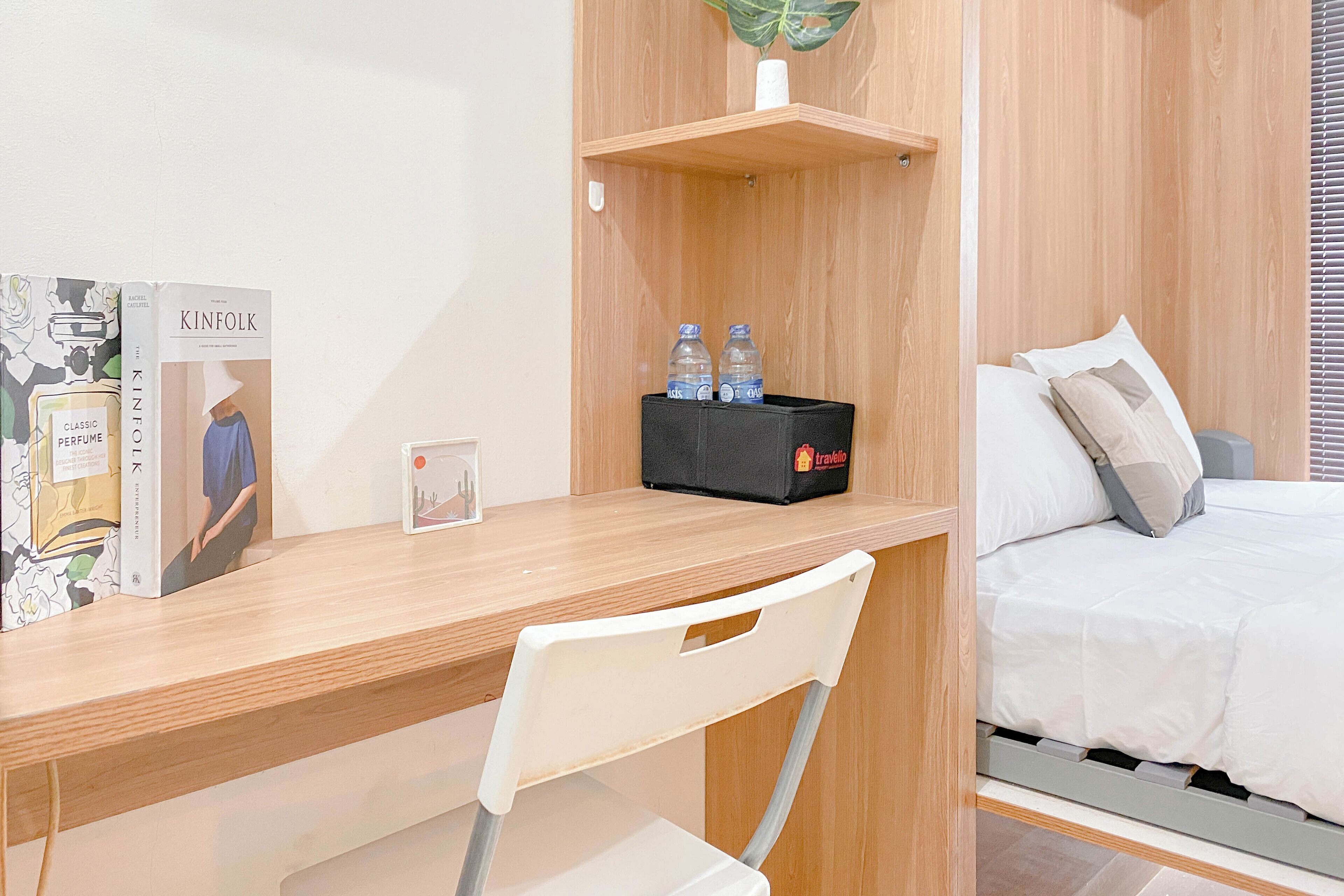 Ein Hotelzimmer mit Holztisch, Stuhl, Bett mit weißer Bettwäsche und einer kleinen Ablage mit zwei Wasserflaschen und einer Taschentuchbox.
