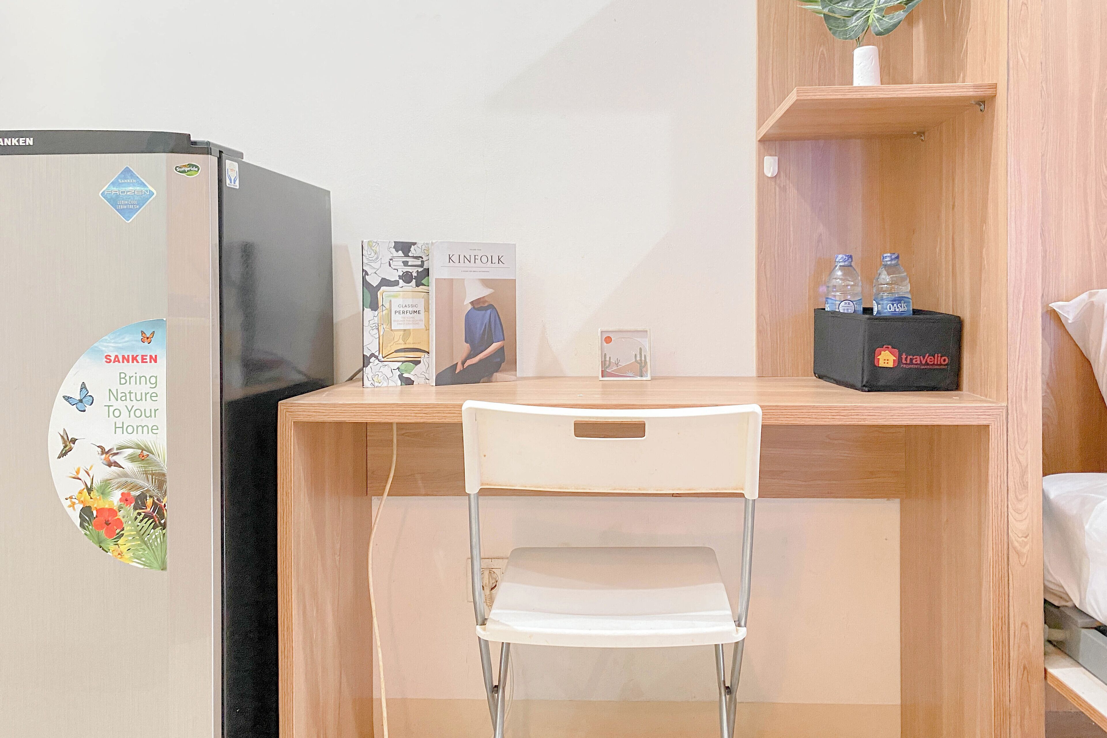 A compact room with a wooden desk, a white chair, a black refrigerator, and a small shelf with water bottles and a toiletry bag.
