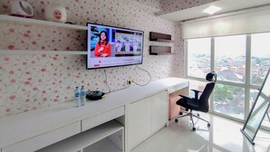 A modern office with a large TV on the wall, a white desk, a black office chair, and a view of the city through large windows. - Comfy and Warm Studio at The Square Surabaya Apartment (Surabaya)