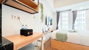A modern hotel room with a wooden desk, a bed with a green blanket, a mounted TV, and a window with curtains. - Cozy and Classical Studio Apartment Tokyo Riverside PIK 2 (Teluknaga)