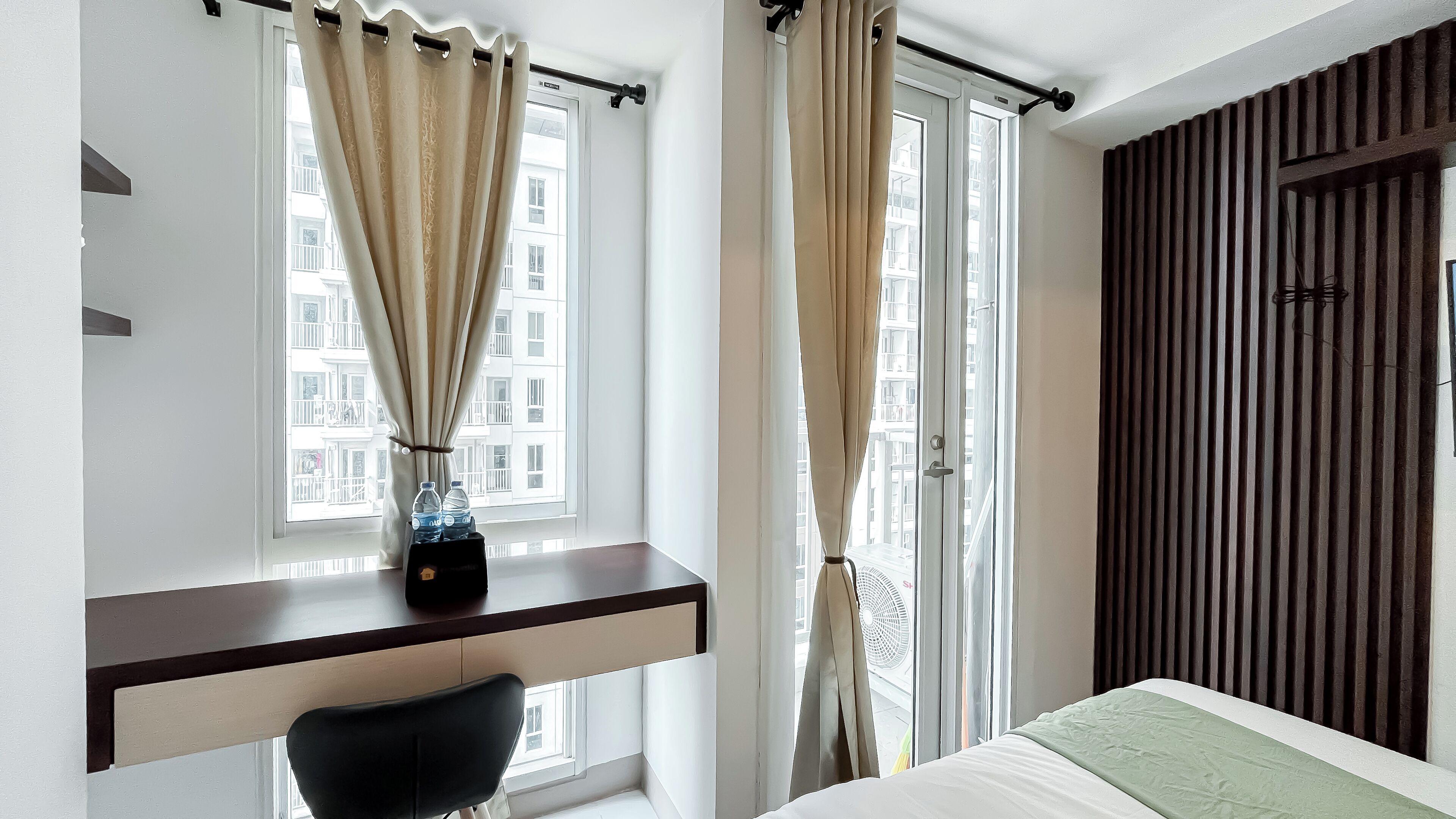 A hotel room with a desk, chair, bed, and a view of the city through large windows.
