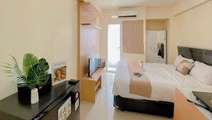 A hotel room with a bed, a TV, a desk with a computer, a plant, and a mirror. - Nice Studio Green Pramuka City Apartment near Shopping Center (Jakarta)