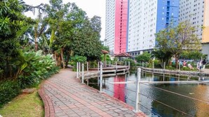 A pathway leading to a small bridge over a water feature, surrounded by lush greenery and tall buildings in the background. - Nice Studio Green Pramuka City Apartment near Shopping Center (Jakarta)