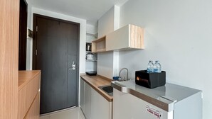 Eine kleine Küche mit einer dunklen Tür, einem Spülbecken, einer Arbeitsplatte und einem Kühlschrank. Auf dem Kühlschrank stehen zwei Wasserflaschen und die Nummer eines Kundencenters ist auf der Kühlschranktür zu sehen.