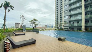 A rooftop pool area with wooden decking, lounge chairs, and a view of a cityscape in the background. - Nice and Comfortable 2BR Apartment at Daan Mogot City (Jakarta)