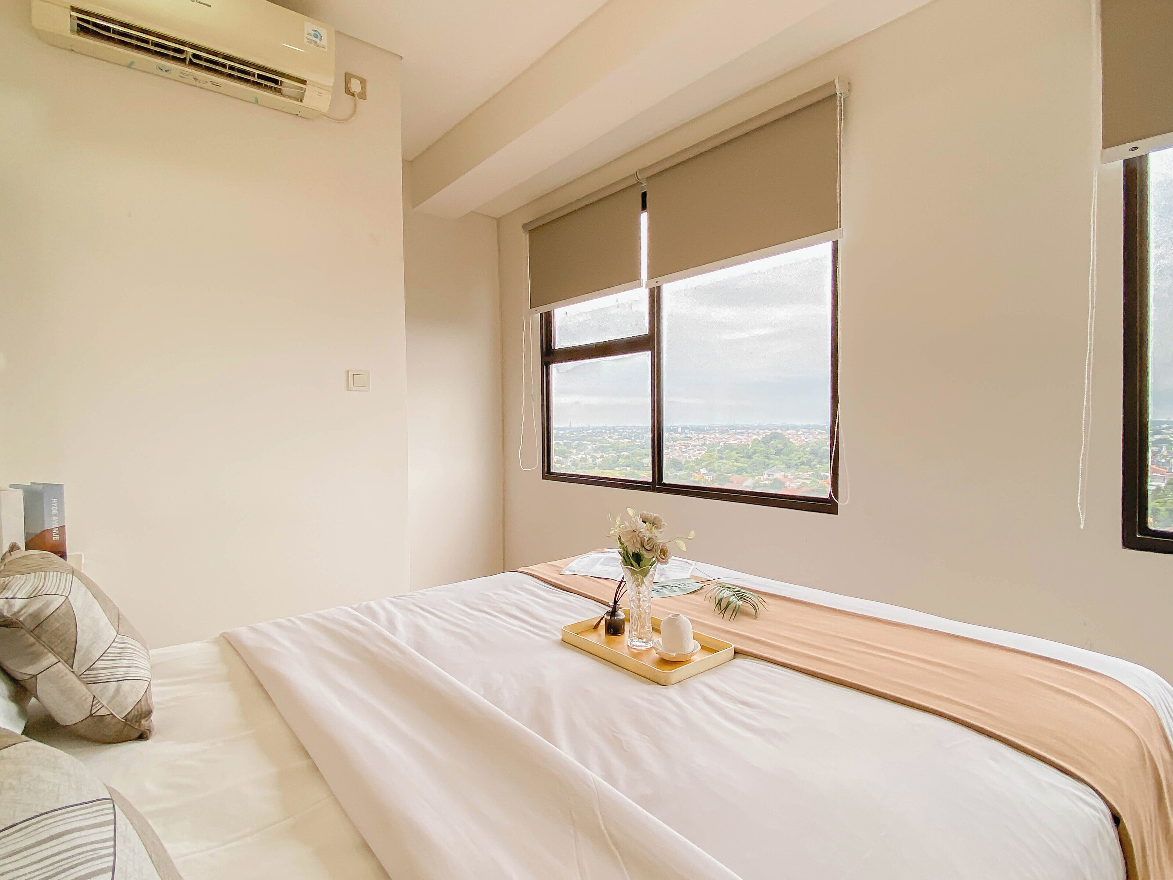 A hotel room with a bed, a window with a view of a cityscape, and an air conditioning unit on the wall.