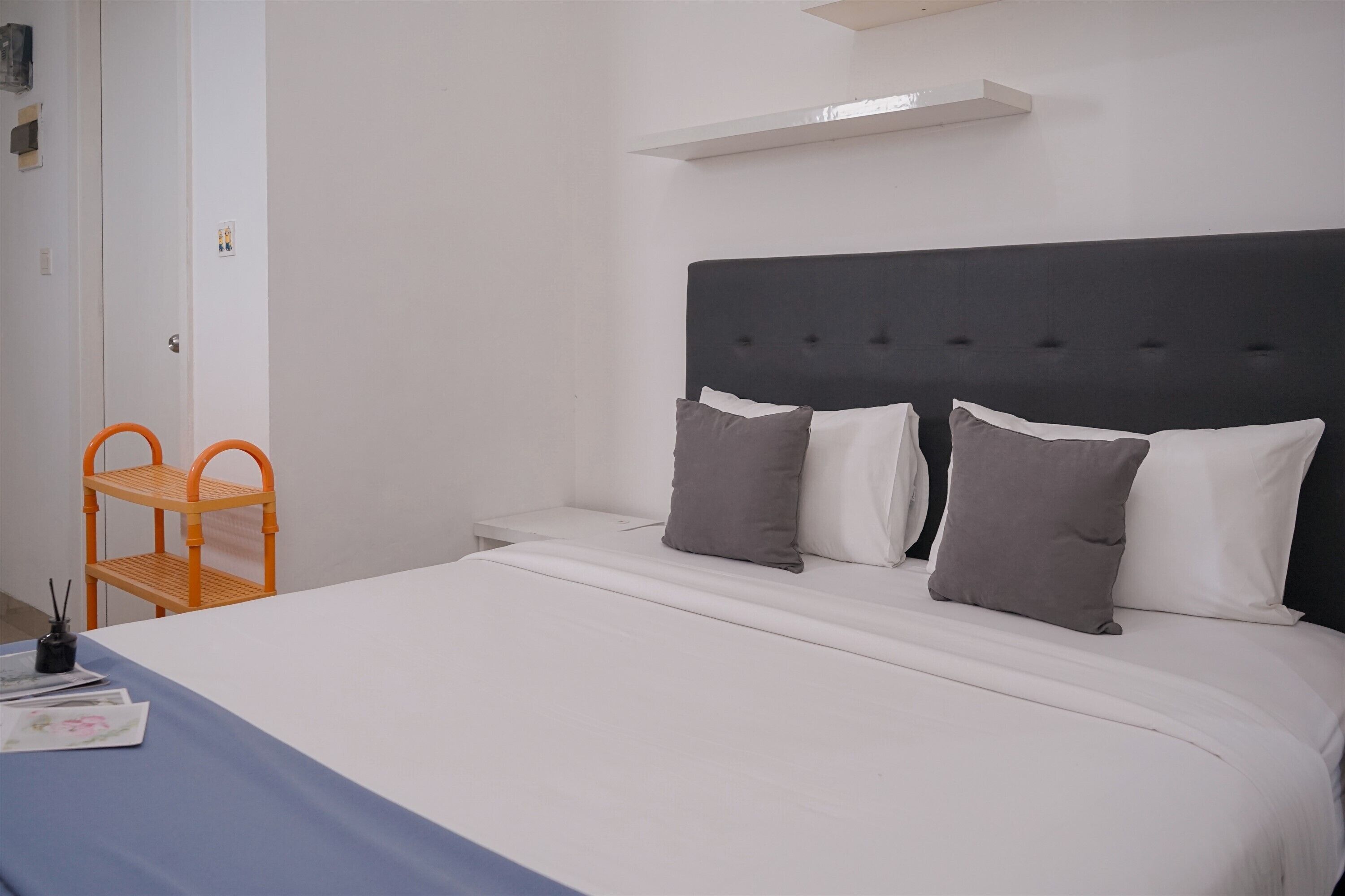 Una cama bien hecha con cabecera oscura, mesita de noche blanca y una silla naranja.