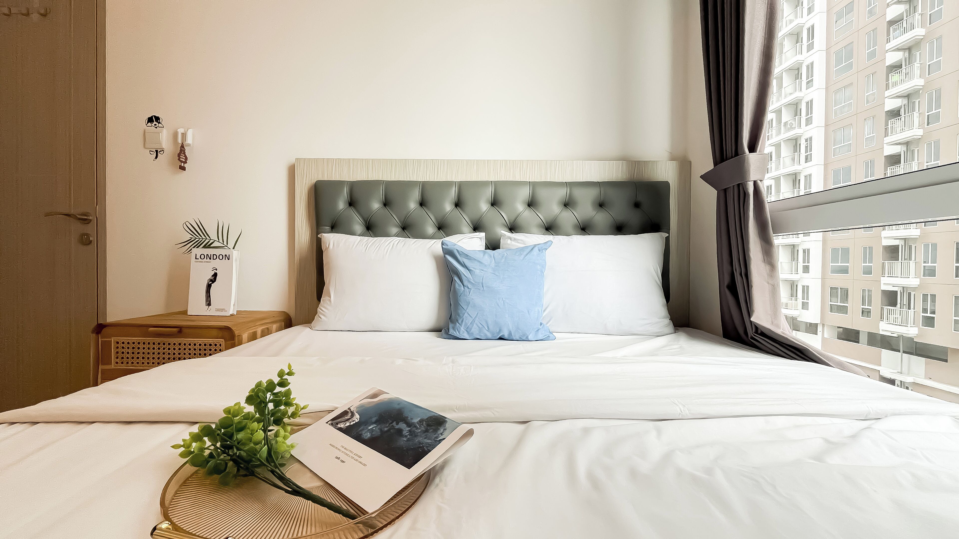Une chambre d’hôtel avec une tête de lit capitonnée, du linge de lit blanc, un oreiller bleu, une table de chevet avec une plante et un livre, et une vue sur un immeuble par la fenêtre.