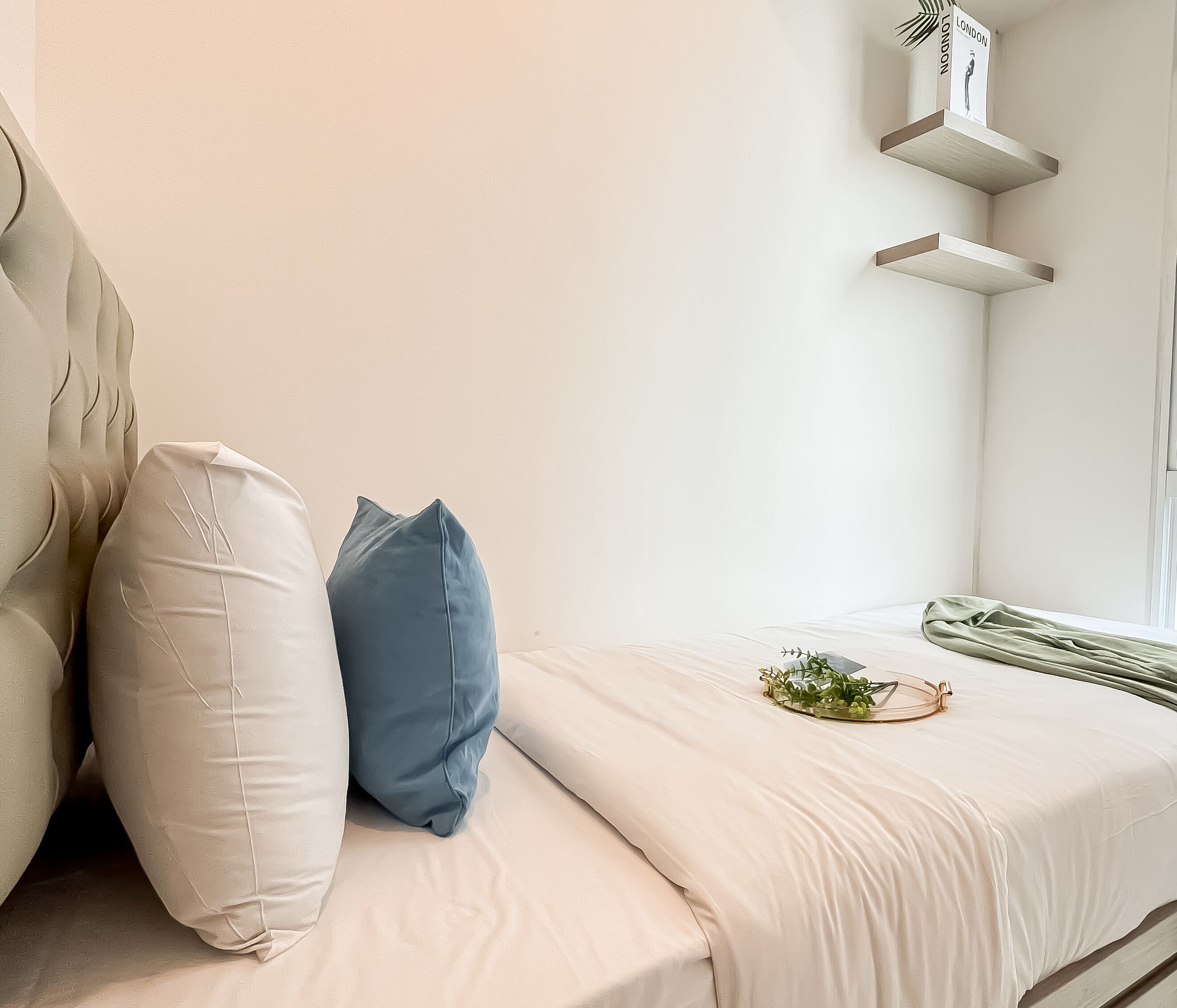 A neatly made bed with a white and blue pillow arrangement, a small plant on the bed, and a shelf with a book titled "LONDON" on the wall.
