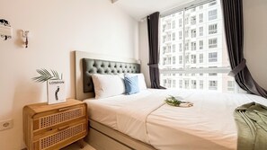 A neatly arranged bedroom with a bed, a bedside table, a wall-mounted light switch, and a window with curtains. - Cozy Design 2BR Apartment Tokyo Riverside PIK 2 (Teluknaga)