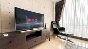 A modern living room with a flat-screen TV displaying 'NETFLIX', a dark wooden entertainment unit, a black leather sofa, and a large window with curtains. - Well Designed 1BR Apartment L'Avenue Pancoran (Jakarta)