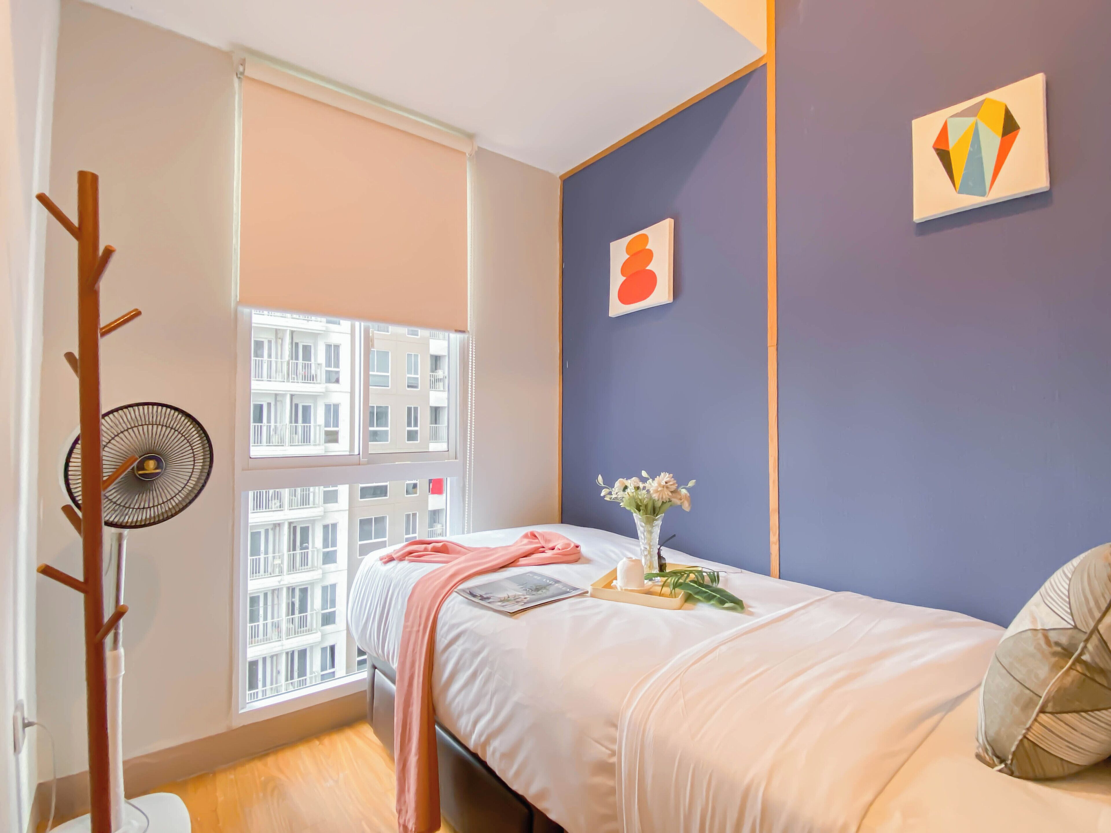 A bedroom with a bed, a window with blinds, a fan, and a vase with flowers.