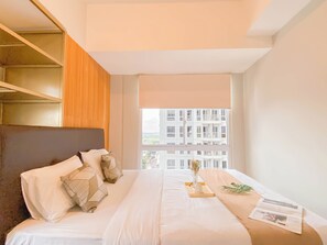 A hotel room with a bed, a wooden headboard, a window with a view of buildings, and a tray with a vase and a book.