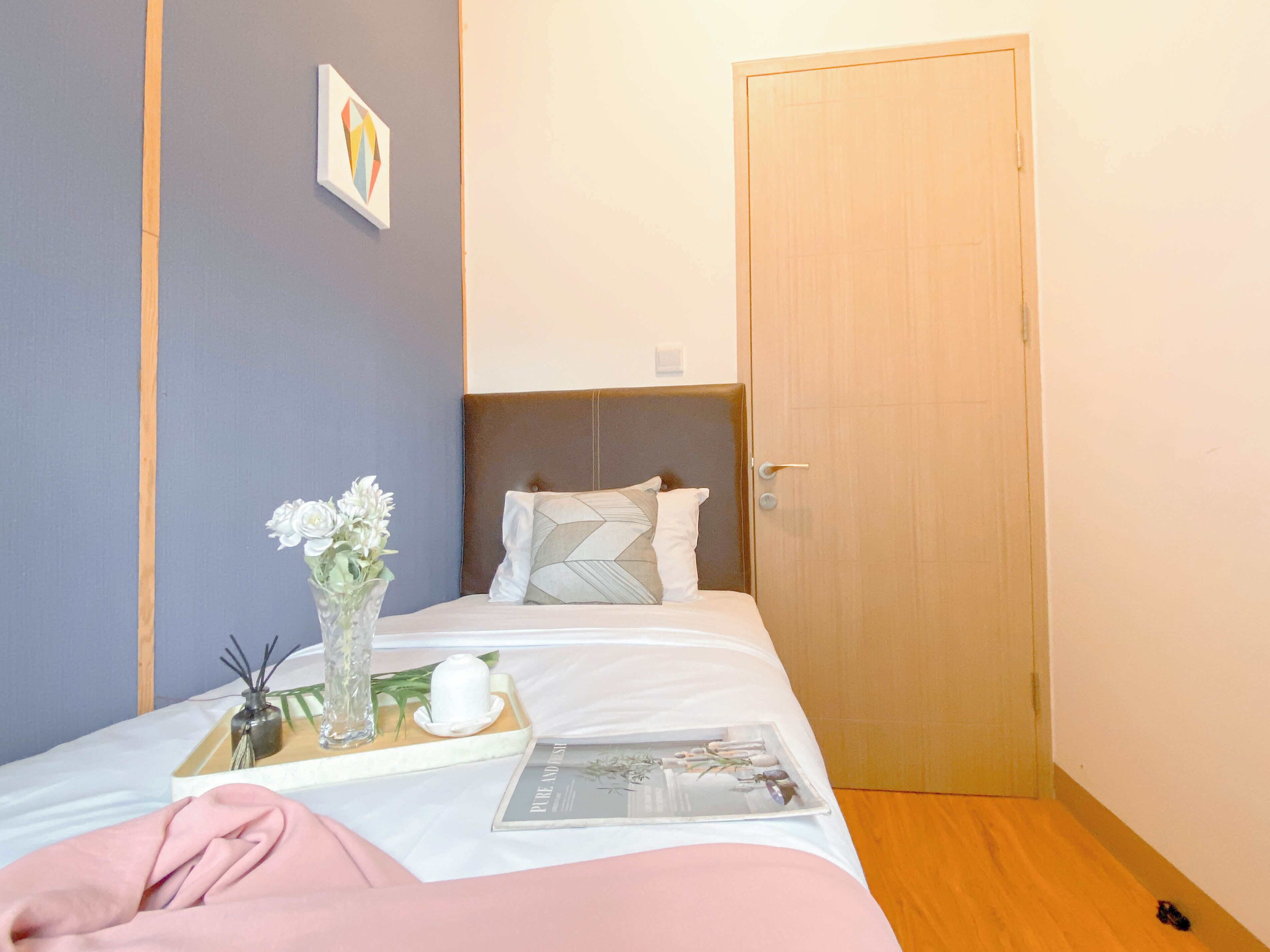 A neatly made bed with a white comforter and pillows, a bedside table with a vase of flowers, a tray with a teapot and a book, and a wooden door in the background.
