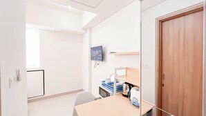 Une pièce avec une porte en bois, un bureau avec un ordinateur et une imprimante, un téléviseur fixé au mur et une fenêtre laissant entrer la lumière naturelle.